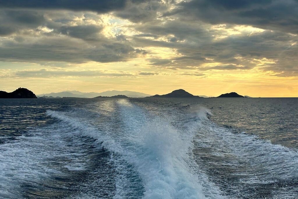 View from a boat leaving a foamy wake in the ocean, with distant islands under a dramatic, cloudy sunset sky.
