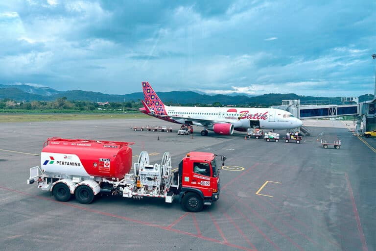 A Pertamina fuel truck is parked on the Labuan Bajo airport tarmac near a Barbie-themed airplane at a gate, with mountains and a cloudy sky in the background.