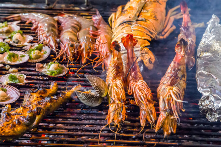 Various seafood, including large prawns, lobster, scallops, and clams, are being grilled over open flames on a barbecue grill. There is also some food wrapped in foil on the right side. Steam and smoke are visible.