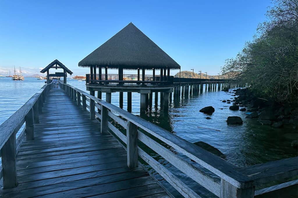 A wooden boardwalk in Labuan Bajo leads over the water to a thatched-roof pavilion on stilts, surrounded by calm blue water and lush green foliage under a clear sky.