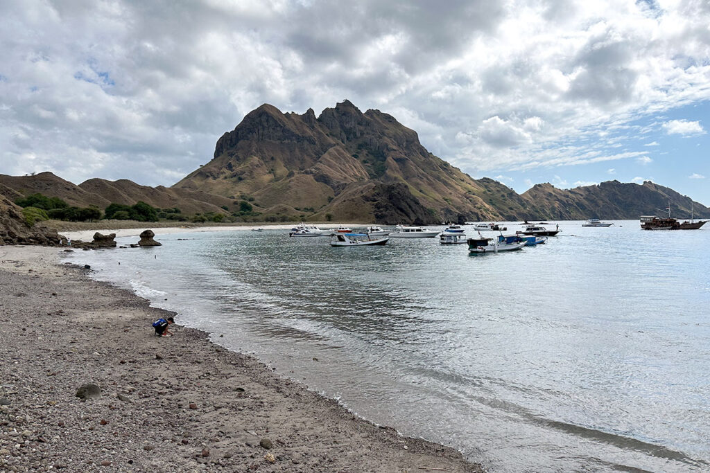A rocky beach with calm water, several small boats floating near the shore, and a rugged mountain rising in the background under a partly cloudy sky.