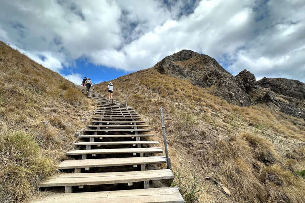 Two people climb a wooden staircase on a dry, grassy hillside under a partly cloudy sky, with the rocky mountain peak of Padar Island near Labuan Bajo in the background.