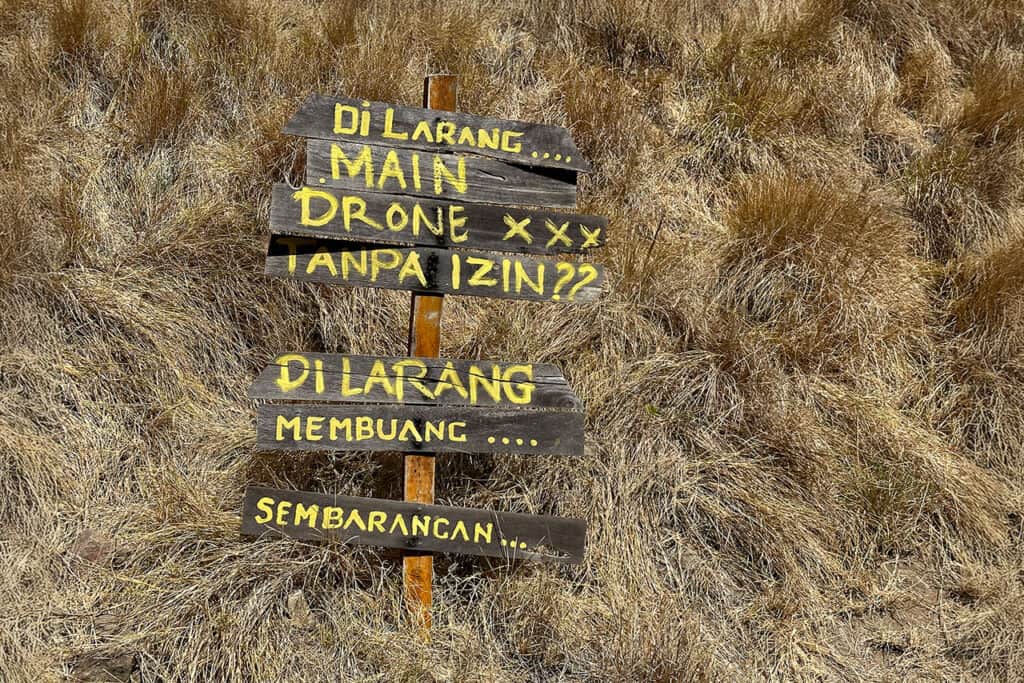 A wooden sign in dry, grassy terrain near Labuan Bajo displays warnings in Indonesian, stating that flying drones and littering are prohibited without permission.