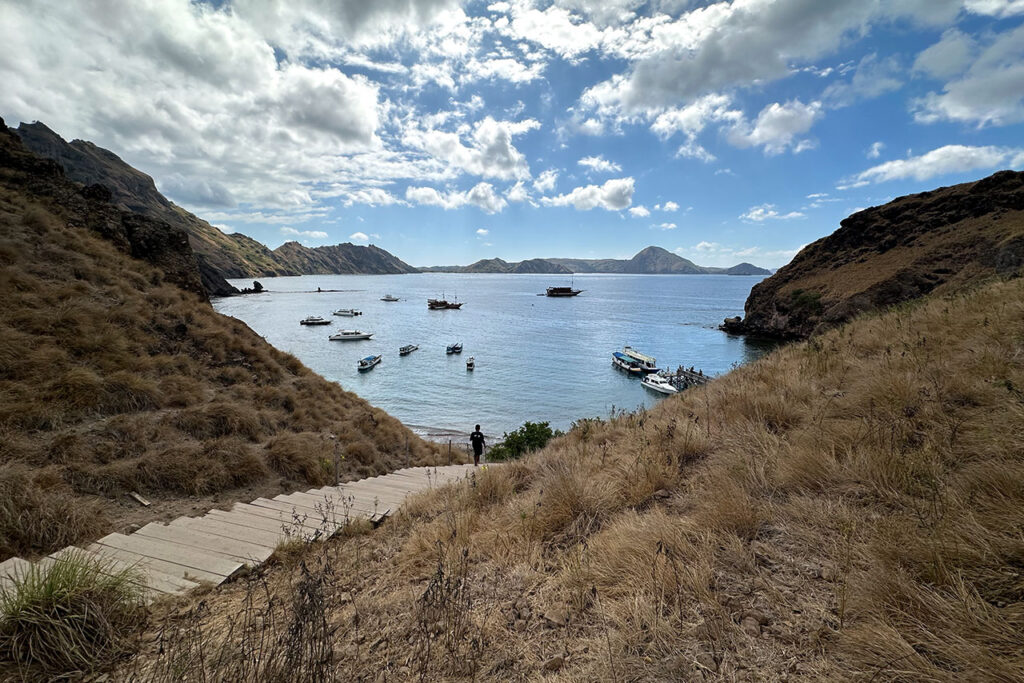 A winding wooden path on a dry, grassy hillside leads to Labuan Bajo’s bay, where boats rest in calm blue waters surrounded by rugged hills. A person stands near the path, gazing over this scenic haven beneath a partly cloudy sky.