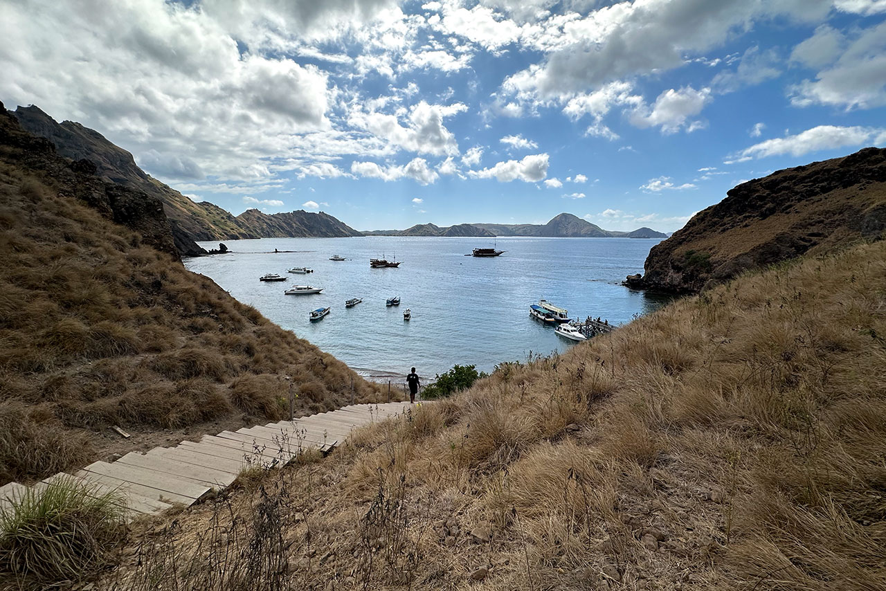 A winding wooden path on a dry, grassy hillside leads to Labuan Bajo’s bay, where boats rest in calm blue waters surrounded by rugged hills. A person stands near the path, gazing over this scenic haven beneath a partly cloudy sky.