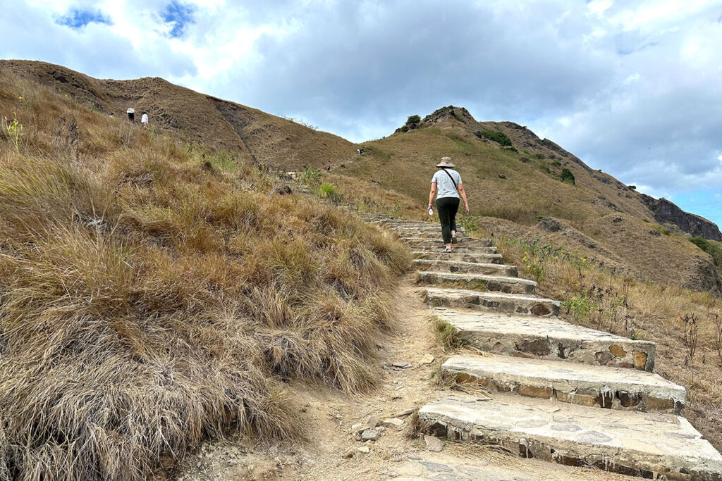 A person wearing a hat and backpack walks up stone steps on a dry, grassy hillside under a cloudy sky, capturing the rugged beauty of Labuan Bajo Padar Island.