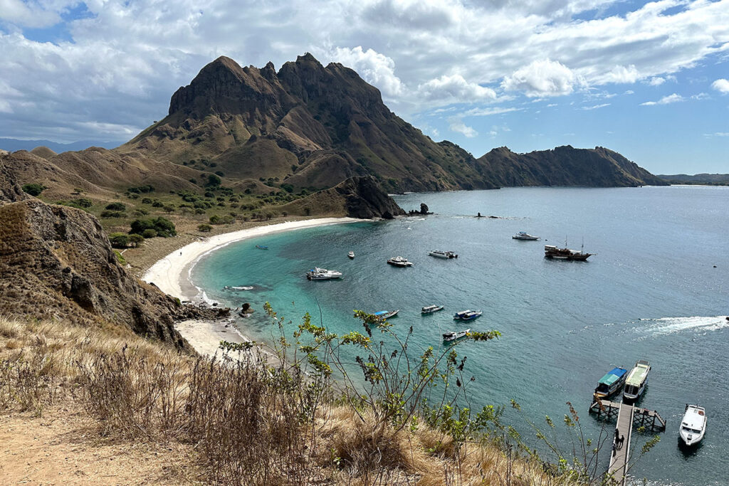 A scenic bay near Labuan Bajo, with clear blue water, several boats anchored off the white sand beach, and the rugged, grassy heights of Padar Island rising in the background under a partly cloudy sky. A scenic bay near Labuan Bajo, with clear blue water, several boats anchored off the white sand beach, and the rugged, grassy heights of Padar Island rising in the background under a partly cloudy sky.