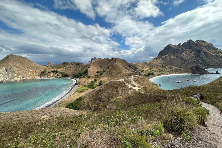 A scenic view of Labuan Bajo’s Padar Island reveals grassy hills, two turquoise bays, and boats anchored near the shore under a partly cloudy sky. A winding path leads along the picturesque hillsides.