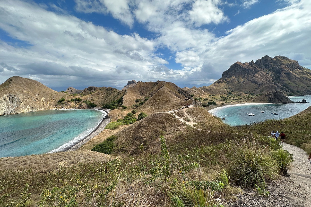 A scenic view of Labuan Bajo’s Padar Island reveals grassy hills, two turquoise bays, and boats anchored near the shore under a partly cloudy sky. A winding path leads along the picturesque hillsides.