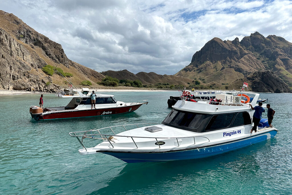 Two motorboats float on clear turquoise water near the rocky shoreline of Labuan Bajo Padar Island, with mountains in the background; a few people stand on each boat under a partly cloudy sky.
