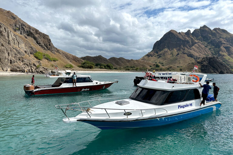 Two motorboats float on clear turquoise water near the rocky shoreline of Labuan Bajo Padar Island, with mountains in the background; a few people stand on each boat under a partly cloudy sky.