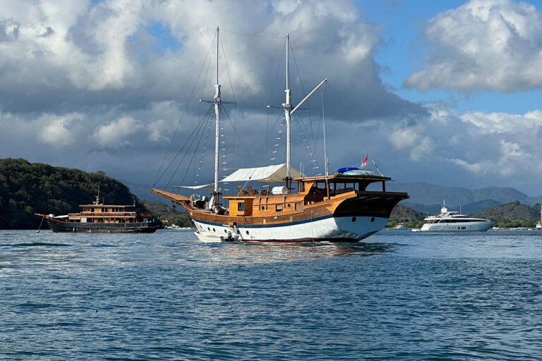 Three boats are anchored on calm blue water: a traditional phinisi boat in the foreground, another wooden boat behind it, and a white yacht in the distance. Forested hills and a cloudy sky form the background.
