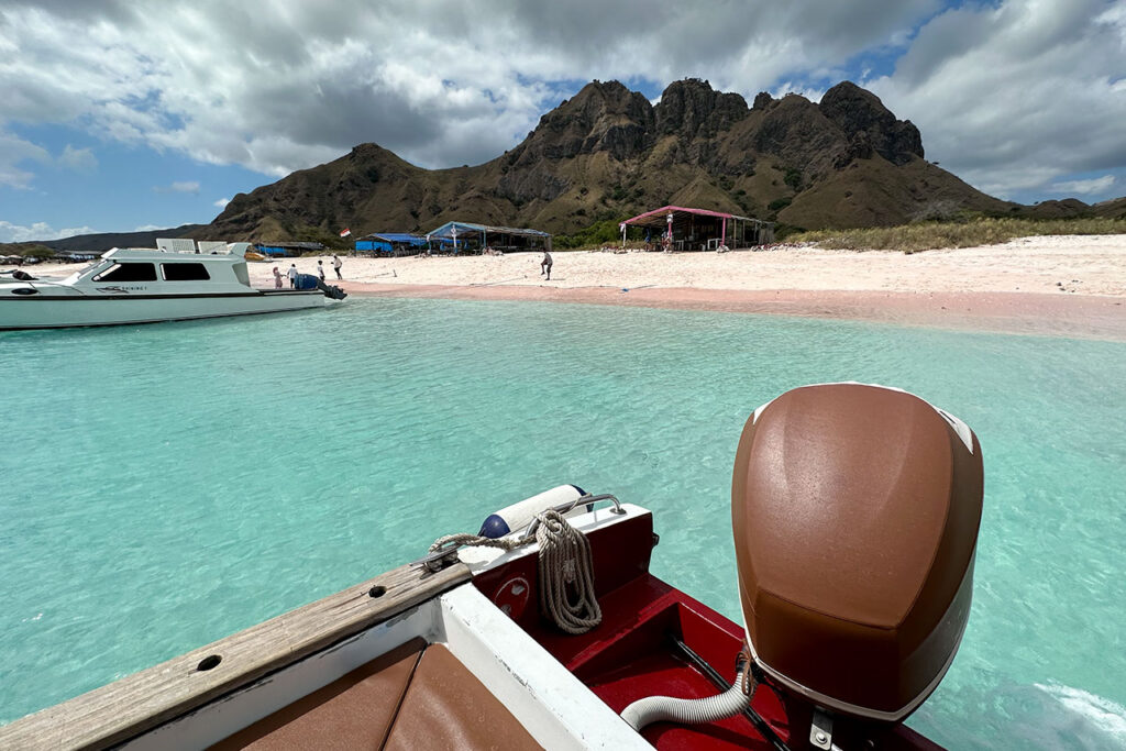 A view from a boat near the shore of Pink Beach Labuan Bajo, with turquoise water, white sand, and people under tents. Rocky hills and partly cloudy skies add to the stunning tropical backdrop. A view from a boat near the shore of Pink Beach Labuan Bajo, with turquoise water, white sand, and people under tents. Rocky hills and partly cloudy skies add to the stunning tropical backdrop.