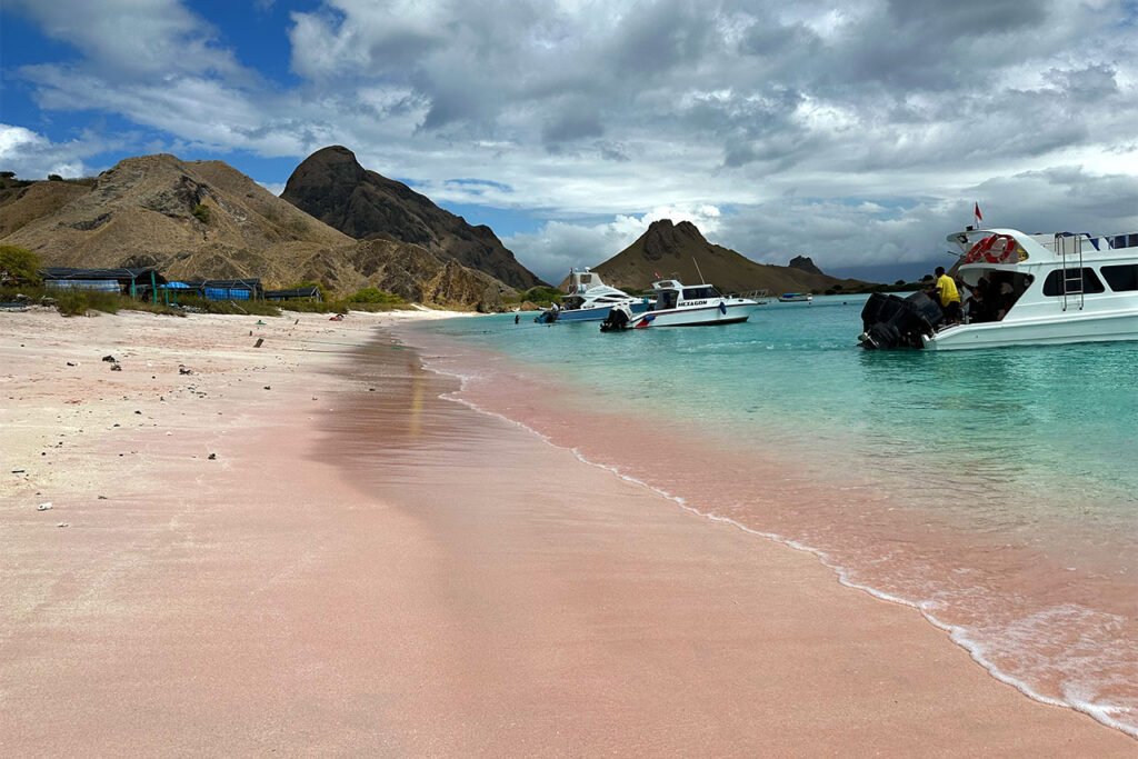 A pink sand beach meets turquoise water at Pink Beach East Nusa Tenggara, with boats docked near the shore. Rocky hills and cloudy skies form the backdrop, while a few people stroll near the boats and along the vibrant shoreline.
