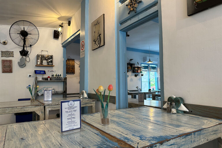 A cozy café and Labuan Bajo restaurant with rustic blue wooden tables, simple decor, a wall-mounted fan, and natural light streaming through windows. A small vase with tulips and a menu sit on the nearest table, with nautical decorations on the walls.