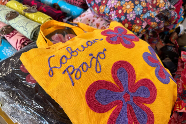 A bright yellow tote bag embroidered with “Labuan Bajo” and large purple and red flowers sits on a market stall in Labuan Bajo, surrounded by colorful patterned fabrics.