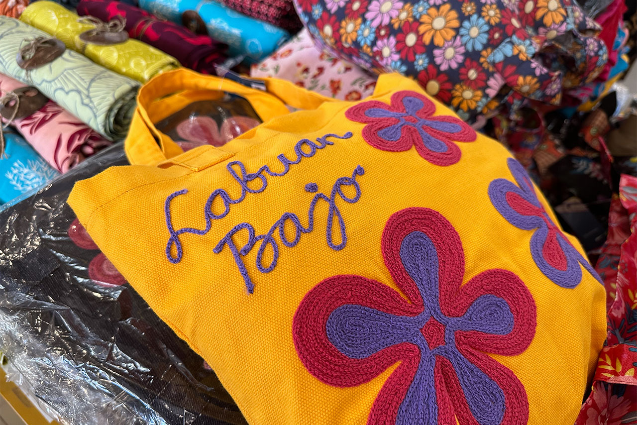 A bright yellow tote bag embroidered with “Labuan Bajo” and large purple and red flowers sits on a market stall in Labuan Bajo, surrounded by colorful patterned fabrics.