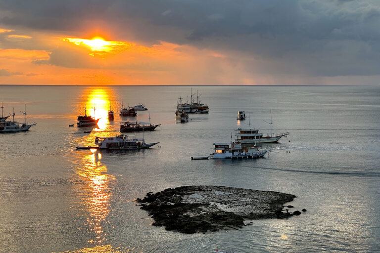 Several boats are anchored on calm water at sunset near Labuan Bajo, with the sun partially hidden by clouds and casting a golden reflection on the sea. A small rocky island is visible in the foreground.