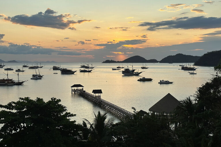 Sunset view over calm water in Labuan Bajo, with boats anchored near a long wooden pier and small gazebos. Silhouetted trees and distant islands appear under a partly cloudy sky.