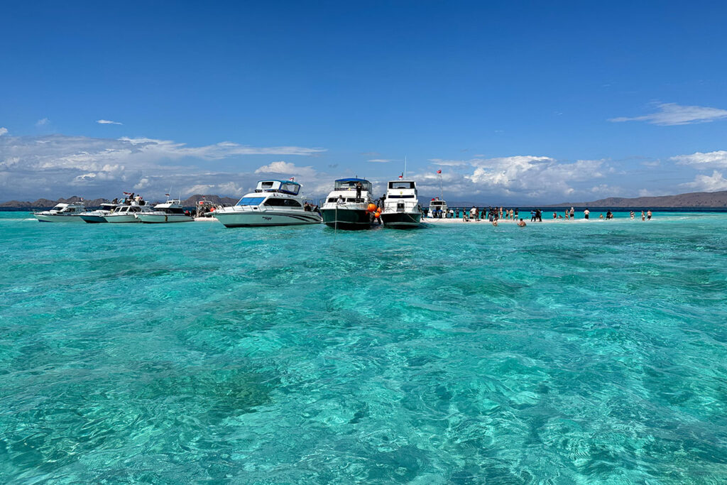 Several boats are anchored in the clear turquoise waters of Taka Makassar, with groups of people swimming nearby under a sunny blue sky and distant mountains in the background.
