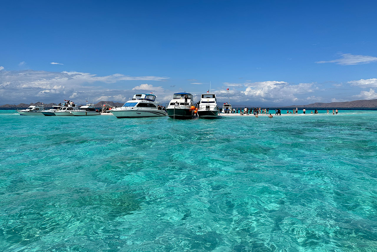 Several boats are anchored in the clear turquoise waters of Taka Makassar, with groups of people swimming nearby under a sunny blue sky and distant mountains in the background.