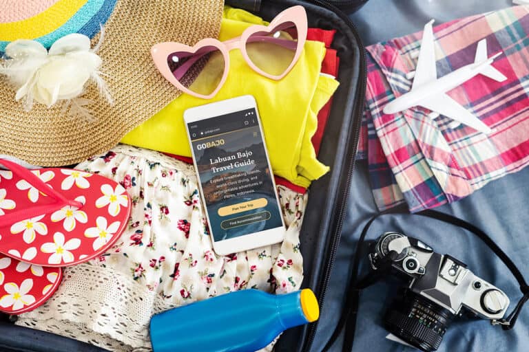 An open suitcase contains clothing, a straw hat, pink heart shaped sunglasses, red flip flops, a water bottle, a camera, a toy airplane, and a smartphone displaying a travel guide.