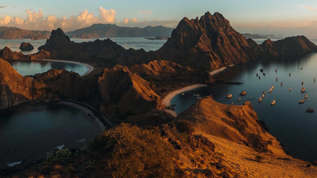 Panoramic view of a rugged coastline with steep hills, curved sandy beaches, clear blue water, and boats anchored near shore at sunset, casting a golden light over the landscape.