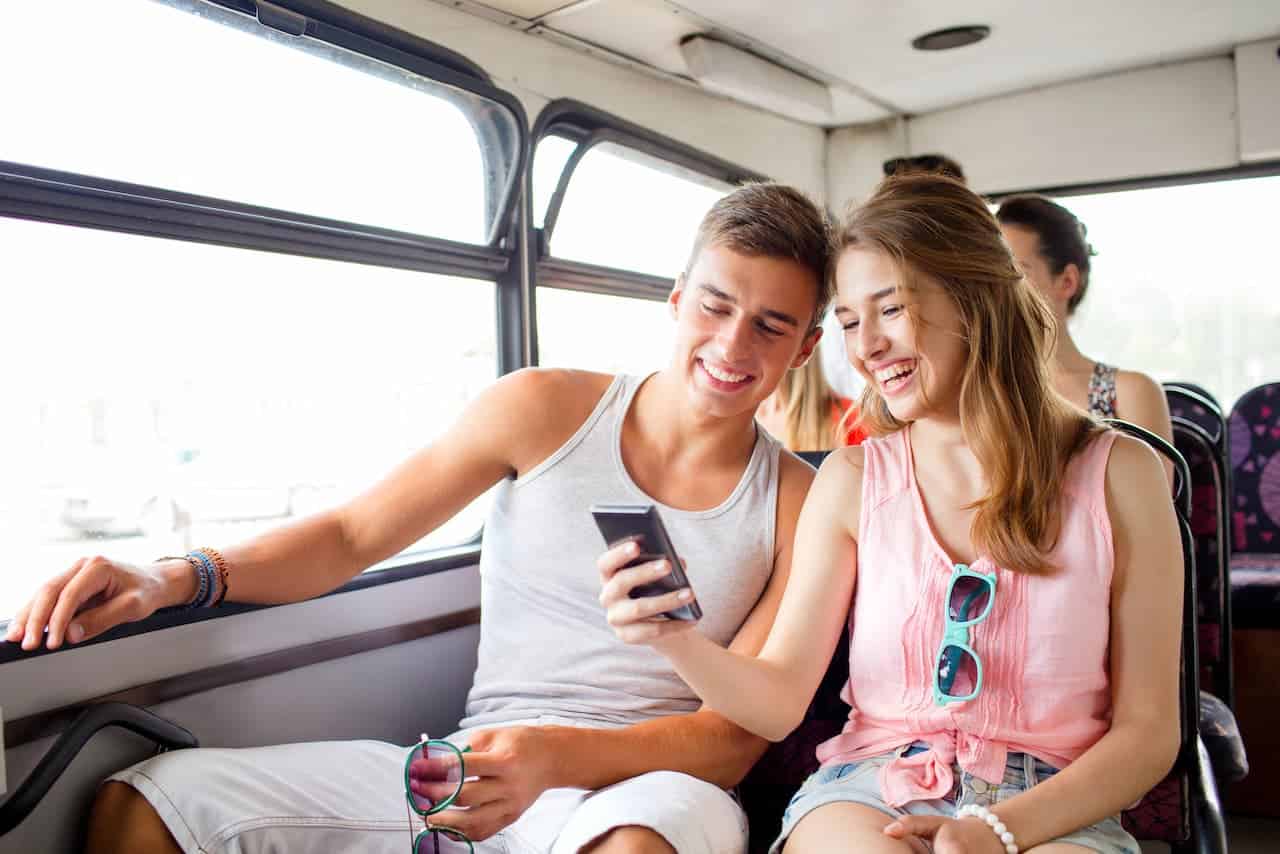 Two smiling young adults sit next to each other on a bus, looking at a smartphone together. Sunlight streams in through the windows, and other passengers are visible in the background.